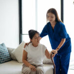 A caregiver in medical scrubs supports an older woman as she rises from the couch. The patient holds a walking aid while receiving guidance. The image highlights home care, rehabilitation support, and safe movement in a home environment.