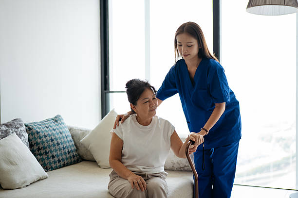 A caregiver in medical scrubs supports an older woman as she rises from the couch. The patient holds a walking aid while receiving guidance. The image highlights home care, rehabilitation support, and safe movement in a home environment.